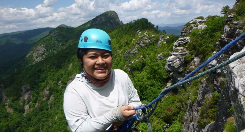 a student pauses to smile at the camera while rock climbing on an outdoor leadership course in the blue ridge mountains. They are wearing safety gear and are secured by ropes.